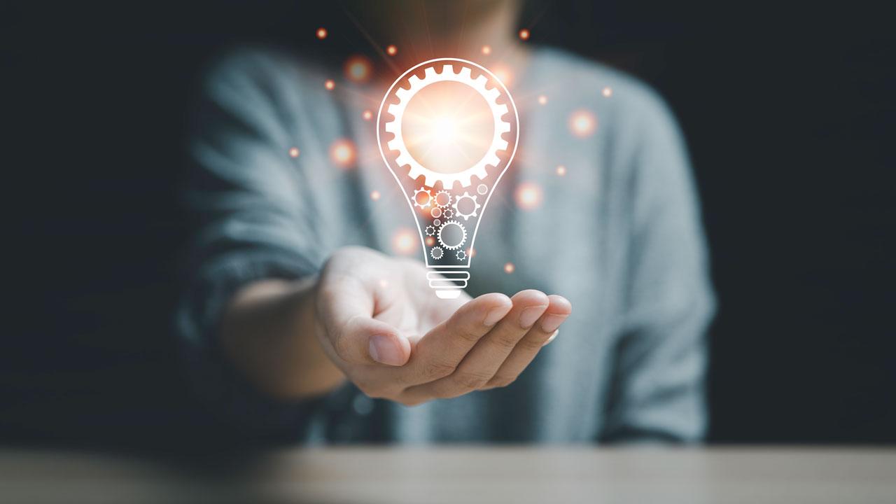 A digital rendering of a lightbulb with gears in glowing light gold floats above the outstretched, cupped hand of a person in a dark room